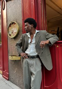 Une personne s'appuie contre un mur, portant un blazer gris sur une chemise à col rayée, un pantalon gris ample et tenant des lunettes de soleil. Fond rouge.