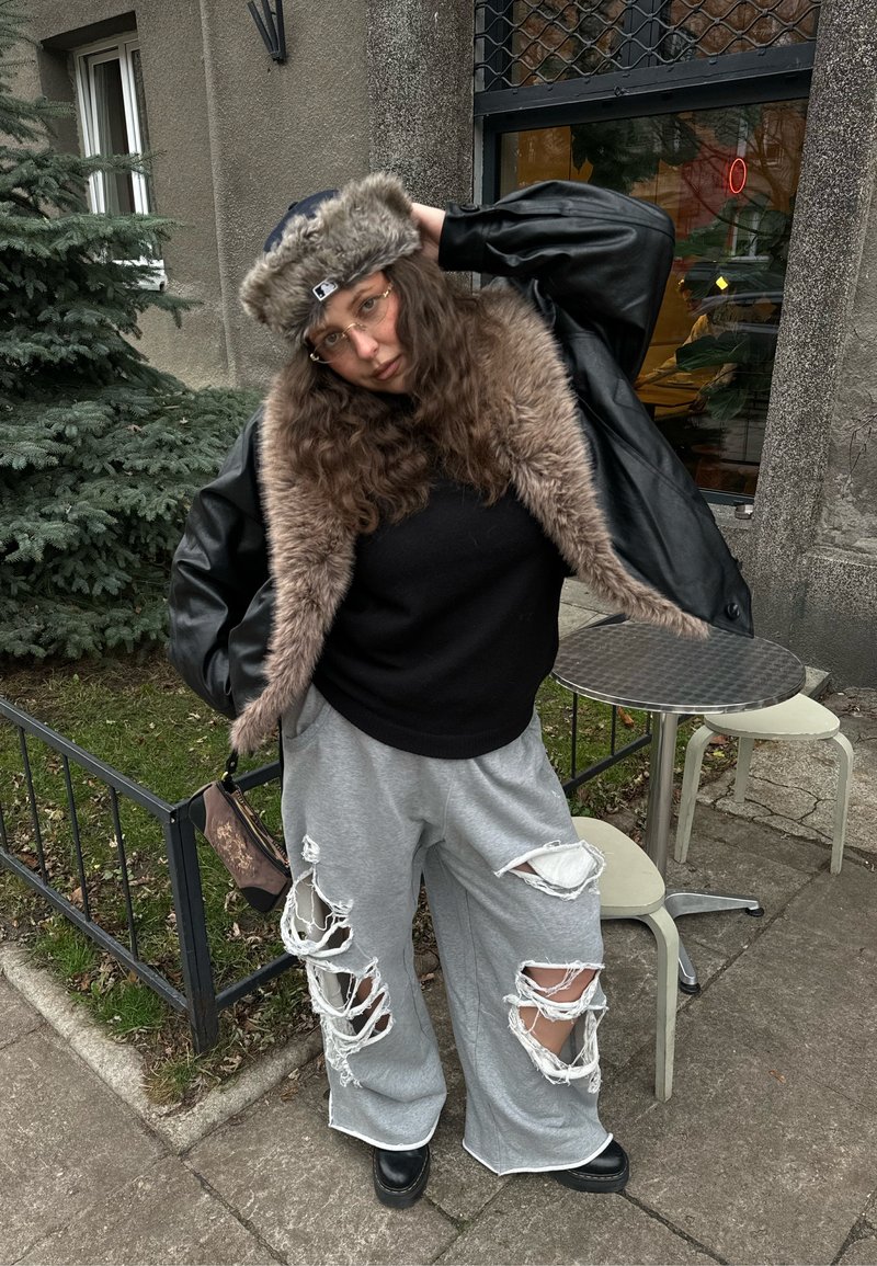 Person mit langen lockigen Haaren, der eine lederne Jacke mit Fellbesatz, einen schwarzen Pullover, zerissene graue Hosen, eine Brille und eine Wintermütze trägt und draußen steht.