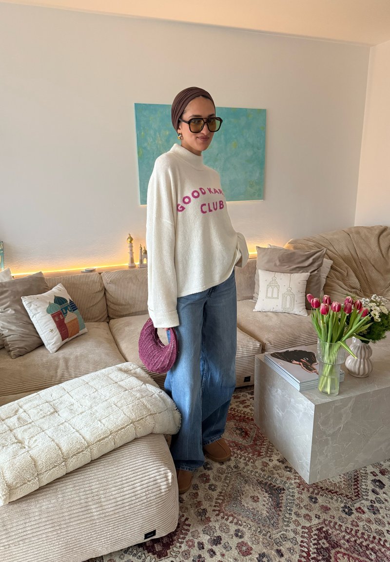 Femme portant un pull blanc "Good Karma Club", un jean bleu, un foulard marron et des lunettes de soleil, debout à côté d'un canapé beige dans un salon chaleureux.