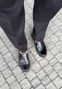 Czarne loafersy z gładkiej skóry, szeroki pasek na wierzchu, w połączeniu z dopasowanymi czarnymi spodniami, na brukowanej nawierzchni.