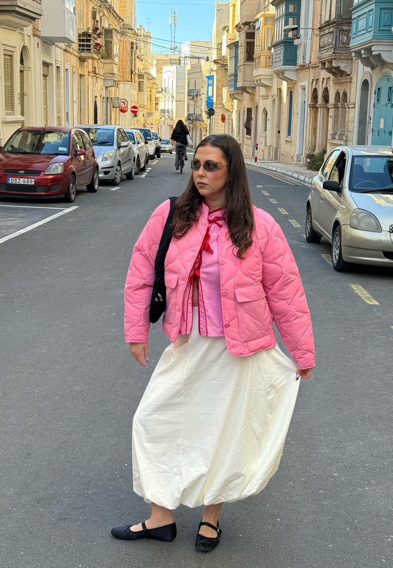 Frau mit pinker Jacke, weißem langem Rock und Sonnenbrille, die auf einer Straße mit parkenden Autos und Gebäuden auf beiden Seiten geht.