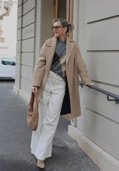 Mujer con abrigo beige, suéter estampado, pantalones blancos de pierna ancha y gafas de sol, sosteniendo un bolso tejido, junto a una barandilla de un edificio al aire libre.