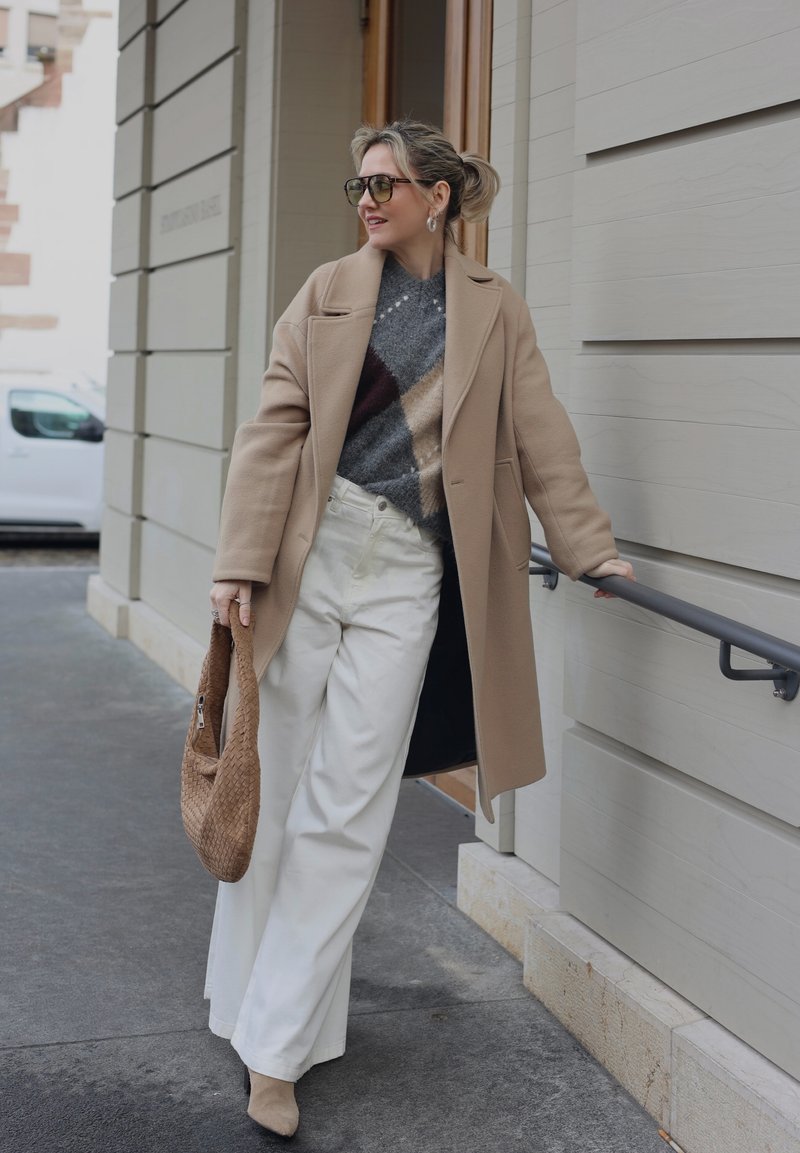 Donna con cappotto beige, maglione a motivo, pantaloni bianchi a gamba larga e occhiali da sole che tiene una borsa intrecciata, in piedi vicino a una ringhiera di un edificio all'aperto.