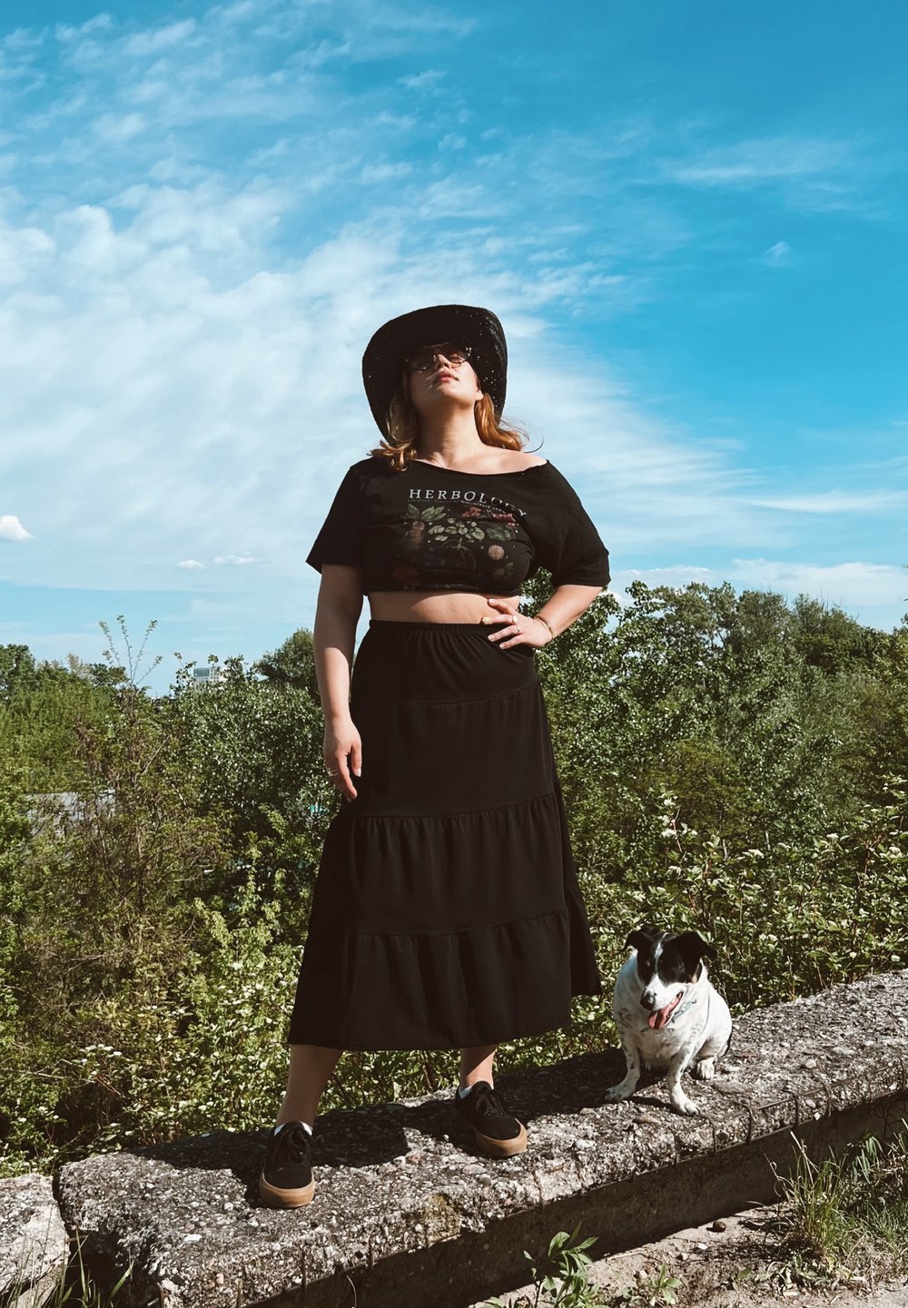 Μαύρο κοντό μπλουζάκι με φλοράλ σχέδιο, μαύρη tiered μακριά φούστα, σκούρα παπούτσια και μαύρο καπέλο με φαρδύ γείσο; σκύλος κάθεται σε μια πέτρινη εξέδρα δίπ�λα.