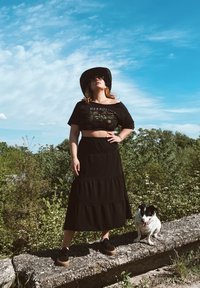 Μαύρο κοντό μπλουζάκι με φλοράλ σχέδιο, μαύρη tiered μακριά φούστα, σκούρα παπούτσια και μαύρο καπέλο με φαρδύ γείσο; σκύλος κάθεται σε μια πέτρινη εξέδρα δίπλα.