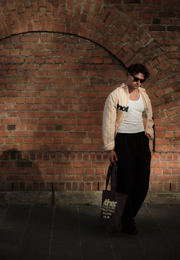 Hombre con gafas de sol, camisa a rayas, camiseta blanca y pantalones negros se apoya en una pared de ladrillos sosteniendo una bolsa oscura.