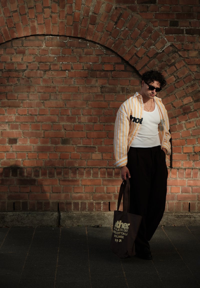 Homme portant des lunettes de soleil, une chemise rayée, un débardeur blanc et un pantalon noir, debout contre un mur de briques, tenant un sac fourre-tout sombre.