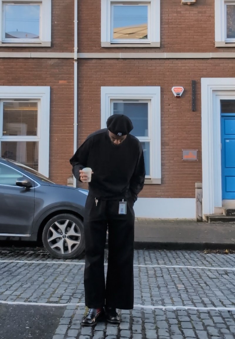 Sweat-shirt noir oversize et pantalon à jambes larges, tenant une tasse de café. Accessoires : un béret et un badge d'identification. Décor de rue urbaine.