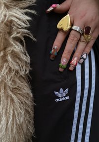 Calças de moletom pretas da Adidas com três riscas brancas. Uma mão exibe unhas longas e coloridas com vários desenhos e anéis, incluindo detalhes em dourado.
