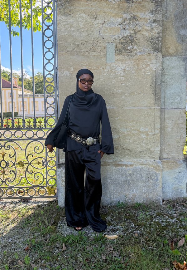 Vrouw met zwarte hijab en zonnebril, gekleed in een zwarte satijnen outfit met een decoratieve riem, staand tegen een stenen muur nabij een sierlijk metalen hek.