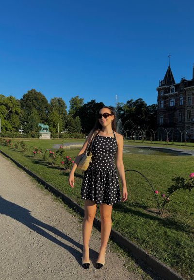 Vestido de lunares blanco y negro con falda acampanada, cuello halter, complementado con un bolso de tejido beige y tacones bicolores negros. Entorno de parque verde.