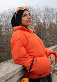 Doudoune orange vif avec de grandes manches, associée à un hijab noir et des lunettes de soleil orange. Cadre extérieur avec des arbres dépouillés.