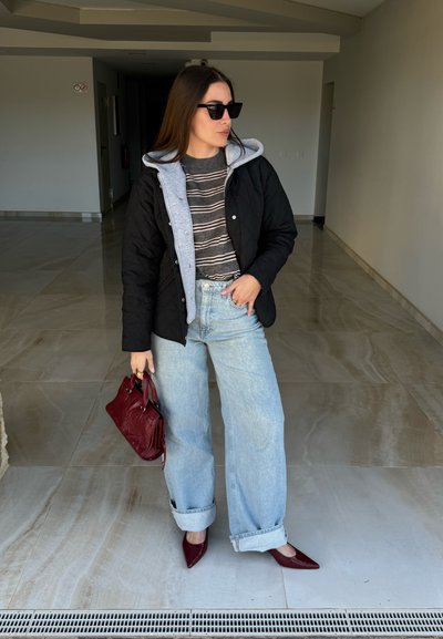 Mujer con cabello largo y castaño, vestida con chaqueta negra, suéter de rayas, pantalones vaqueros anchos de color azul claro, gafas de sol, sosteniendo un bolso rojo, de pie en un pasillo.