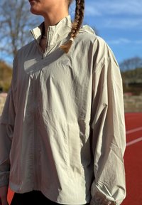 Ljusbeige huvad jacka av texturerat tyg, med blixtlås fram, sidofickor och elastiska muddar. Synligt märke på bröstet.