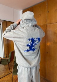 Personne portant un sweat à capuche gris avec une lettre "P" stylisée en bleu dans le dos, ajustant la capuche à l'intérieur près d'armoires en bois et d'une chaise verte.