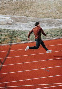Athlète portant un maillot à manches longues bordeaux et un pantalon noir courant sur une piste rouge. Chaussures de sport blanches ; herbe en arrière-plan.