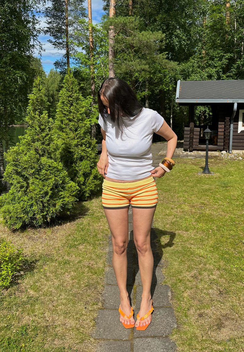 Femme en t-shirt blanc, short rayé jaune et sandales orange debout sur un chemin de jardin, ajustant son short, avec des arbres verts et une cabane en bois à proximité.