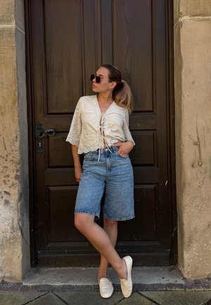 Blusa de eyelet crema con detalle de lazo en el frente, combinada con pantalones cortos bermuda de denim azul y mocasines crema, contra una puerta de madera.