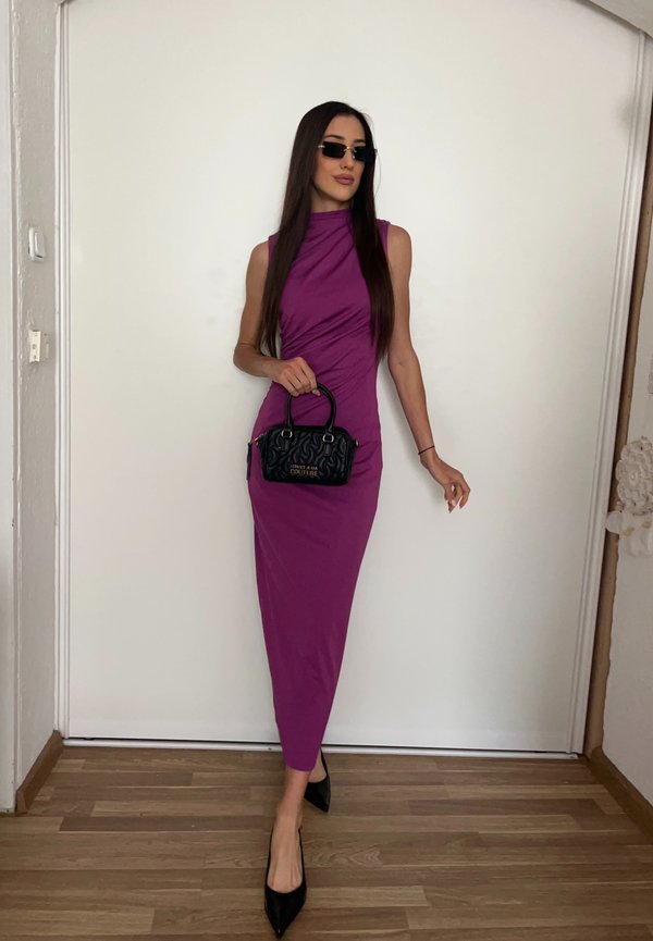 Mujer con un vestido morado sin mangas, tacones negros puntiagudos y gafas de sol, sostiene un pequeño bolso negro, de pie frente a una pared blanca.
