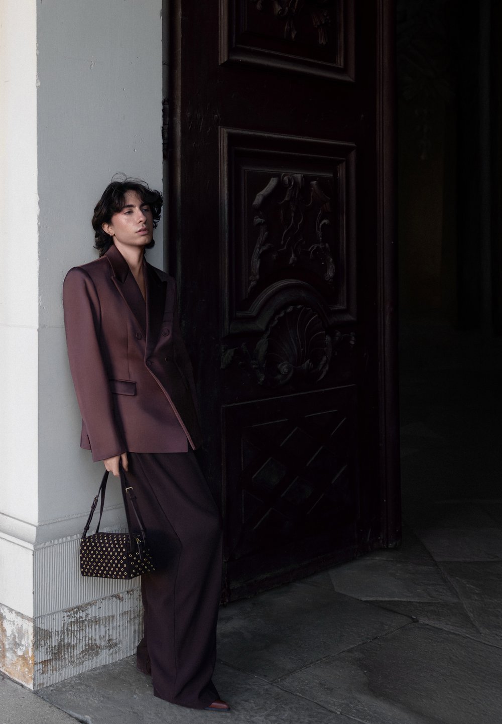Personne en costume oversize violet foncé appuyée contre une colonne blanche, tenant un sac à main noir, debout à côté d'une grande porte en bois sombre ornée.
