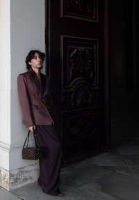 Personne en costume oversize violet foncé appuyée contre une colonne blanche, tenant un sac à main noir, debout à côté d'une grande porte en bois sombre ornée.