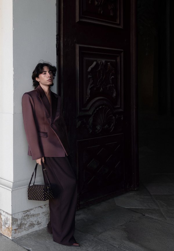 Persona con un ampio completo viola scuro appoggiata a una colonna bianca, che tiene una borsa nera, in piedi accanto a una grande porta in legno scuro ornate.