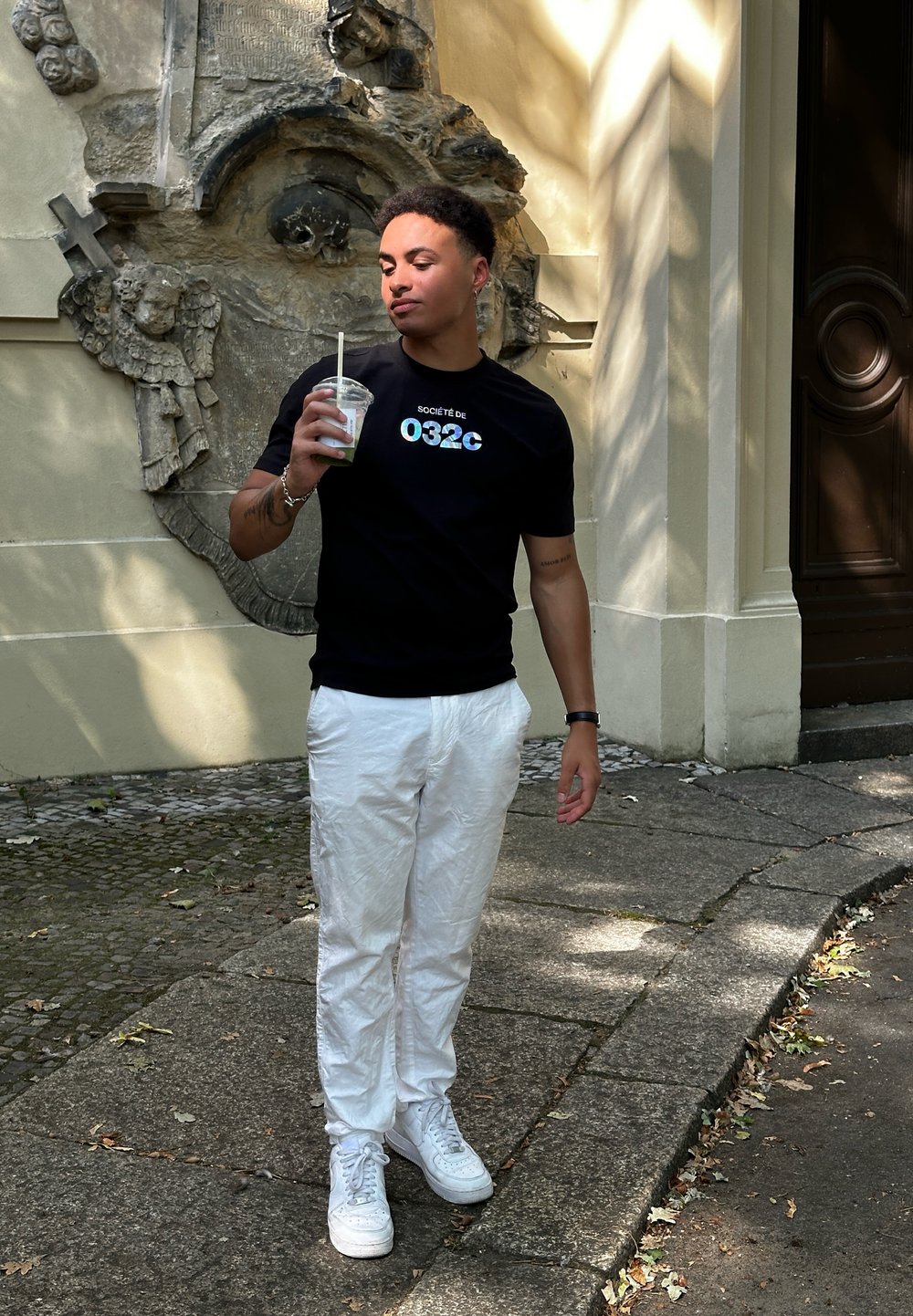 T-shirt noire avec le logo "Société de 032c", pantalon texturé blanc et baskets blanches. Tient un gobelet transparent avec une paille. Fond en pierre.