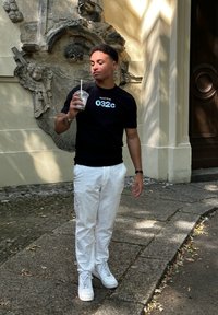 T-shirt noire avec le logo "Société de 032c", pantalon texturé blanc et baskets blanches. Tient un gobelet transparent avec une paille. Fond en pierre.