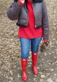 Jachetă maro scurtă cu guler negru de blană artificială, pulover roșu tricotat cu model împletit, blugi albaștri și cizme roșii din cauciuc. Geantă maro cu curea cu lanț.