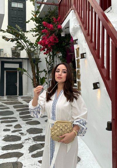 Vestido de lino blanco con bordados azules, mangas abullonadas y un clutch dorado tejido. El fondo presenta paredes blancas, un camino de piedra y vegetación.