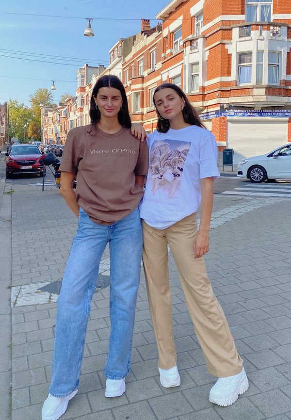 Dos mujeres están en una calle. Una lleva una camiseta marrón y jeans de color azul claro; la otra lleva una camiseta blanca con un gráfico de un lobo y pantalones beis. Ambas tienen zapatillas blancas.