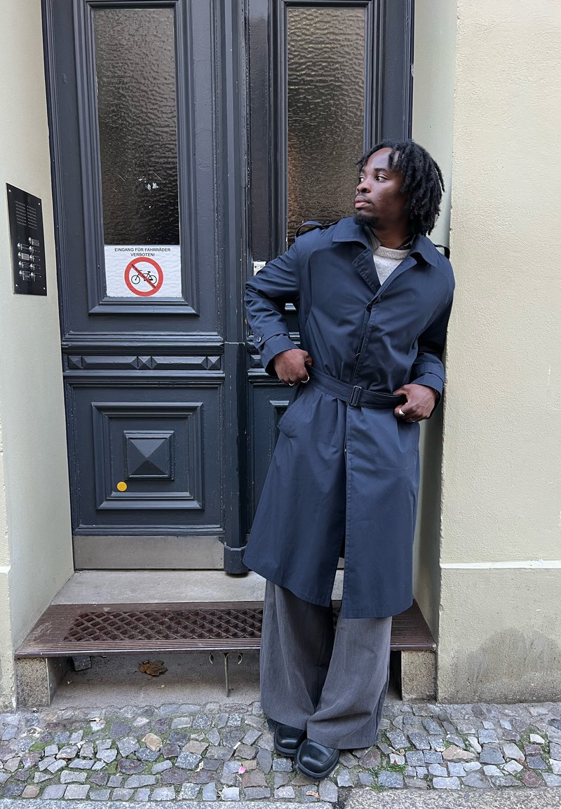 Langer marineblauer Trenchcoat mit Gürtel, Seitentaschen und Schulterklappen. Graue Hosen mit weitem Bein und schwarze Clogs. Im Hintergrund ist eine Tür aus strukturiertem Glas zu sehen.