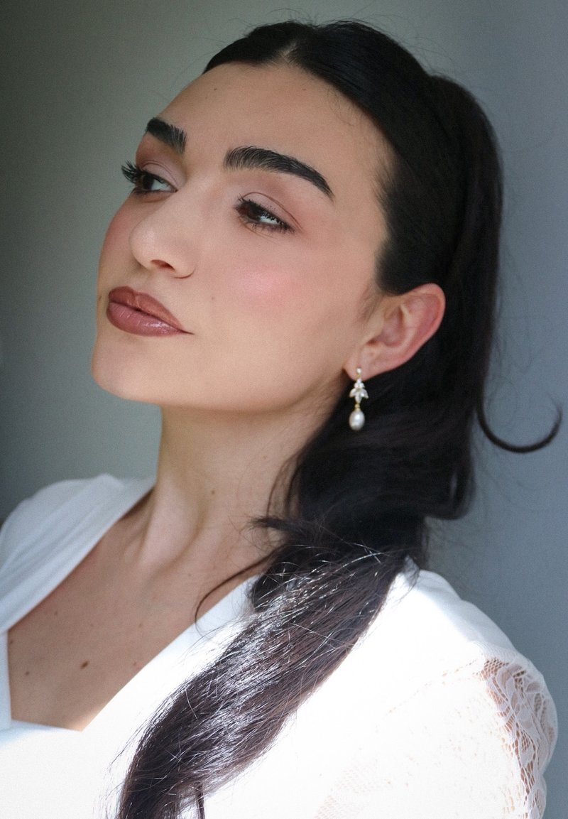 Femme aux longs cheveux noirs attachés, portant des boucles d'oreilles en perles pendantes et un haut blanc à manches en dentelle, regardant calmement sur le côté devant un fond uni.