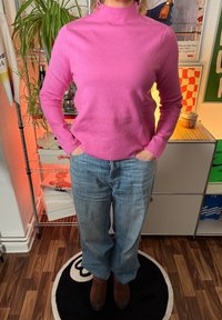 Pulover roz cu guler înalt, textură nervurată, design mulat. Asortat cu blugi albi largi în nuanță de albastru deschis și ghete maro până la gleznă. Fundal cu plante.