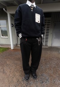 Personne portant un pantalon et des chaussures noirs, un pull noir avec une poche blanche et un col blanc, des colliers superposés et des chaînes, debout en plein air sur du gravier.