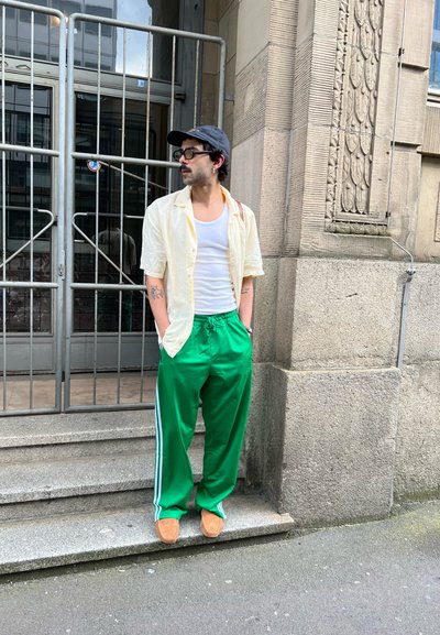 Camisa corta de manga amarilla clara, blusa blanca, pantalones deportivos verdes con raya blanca y zapatos marrones sin cordones. De pie en unos escalones de piedra.