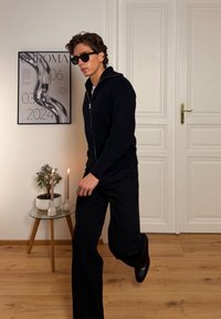 Pull noir à fermeture éclair en tricot, pantalon noir et chaussures noires. Lunettes de soleil et bague portées. Mur blanc et sol en bois avec une plante et des bougies.