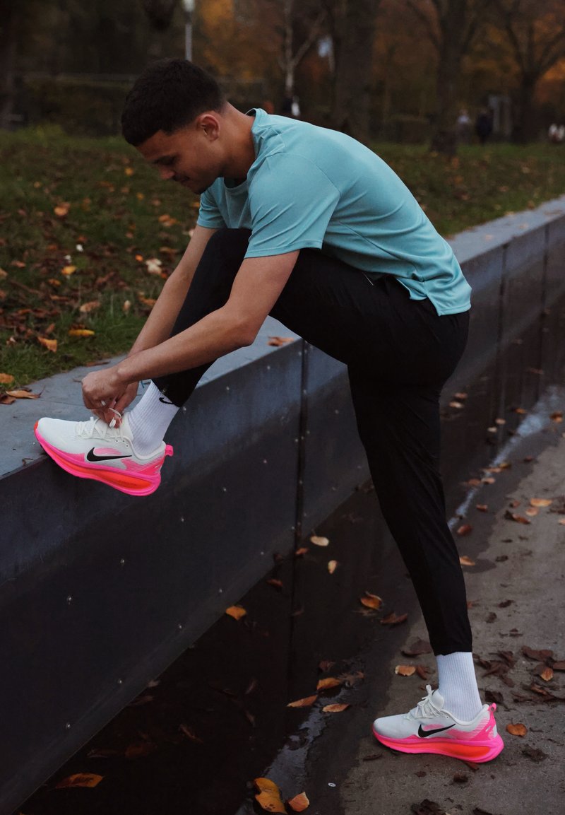 Chaussures de sport Nike blanches avec semelles roses, accents noirs et surface texturée. Une personne est assise, en train de faire ses lacets, portant un pantalon noir.