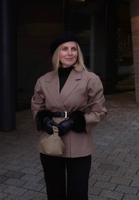 Femme portant un blazer beige avec des poignets en fourrure noire, un béret noir, des gants, et tenant un sac à main beige, debout sur un sol pavé en pierre à l'extérieur.