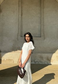 Weißes, strukturiertes, knielanges Kleid mit kurzen Ärmeln; hält eine dunkelbraune geflochtene Handtasche. Der Hintergrund zeigt eine neutralfarbige Wand.