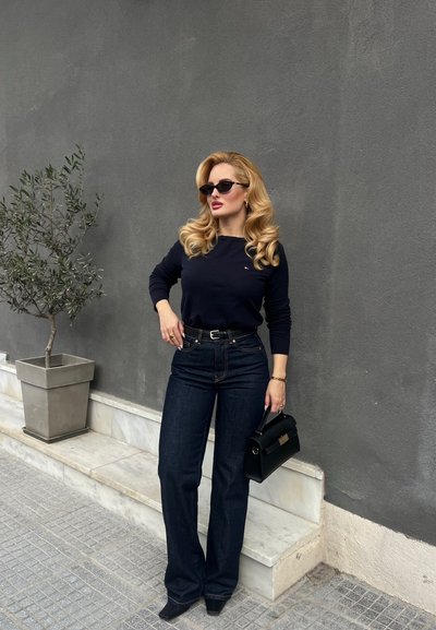 Mujer con rizos rubios que lleva gafas de sol negras, suéter azul marino, jeans oscuros y zapatos de tacón negros, sostiene un bolso negro mientras está de pie junto a una pared gris con una planta en maceta.