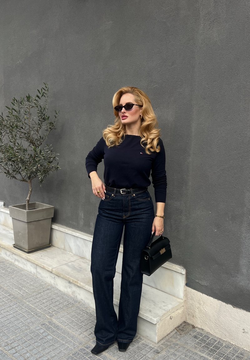 Femme aux boucles blondes portant des lunettes de soleil noires, un pull bleu marine, un jean foncé et des talons noirs, tenant un sac à main noir, debout près d'un mur gris avec une plante en pot.