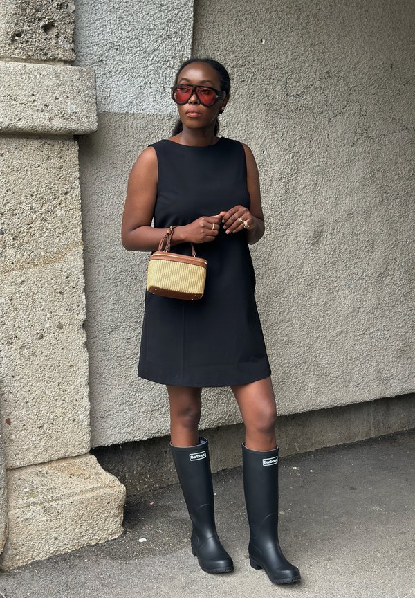 Femme portant une robe noire sans manches, de grandes lunettes de soleil rouges, tenant un petit sac en osier, et des bottes noires Barbour montantes jusqu'au genou.