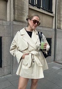 Trench-coat beige avec une ceinture nouée à la taille, grand col et détails à boutons. La personne porte un breuvage vert et des lunettes de soleil surdimensionnées.