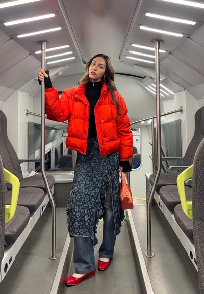 Mujer con chaqueta acolchada roja, falda floral, jeans y zapatos rojos, de pie dentro de un tren moderno y vacío, sujetando un poste y una bolsa naranja.