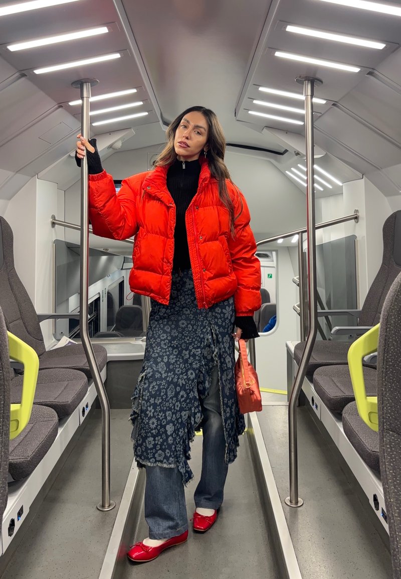 Femme en doudoune rouge, jupe fleurie, jean et chaussures rouges, debout dans un train moderne et vide, tenant une barre et un sac orange.