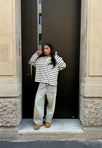 Gestreiftes Langarmshirt, weiß mit schwarzen Linien, kombiniert mit hellblauen weit geschnittenen Jeans und braunen Holzschuhen, stehend auf einer Marmorstufe.