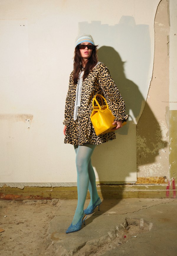 Mujer con vestido de estampado de leopardo, medias azules, tacones azules, gorro de rayas, gafas de sol, sosteniendo un bolso amarillo brillante y de pie en el interior.