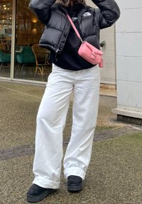Doudoune noire, sweat à capuche noir, pantalon large blanc, sac bandoulière rose et chaussures noires. Cadre extérieur avec un sol texturé.