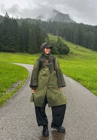 Grønn vanntett regnjakke med hette, brukt over svarte løse bukser og robuste svarte sko. Regndråper synlige på jakken.